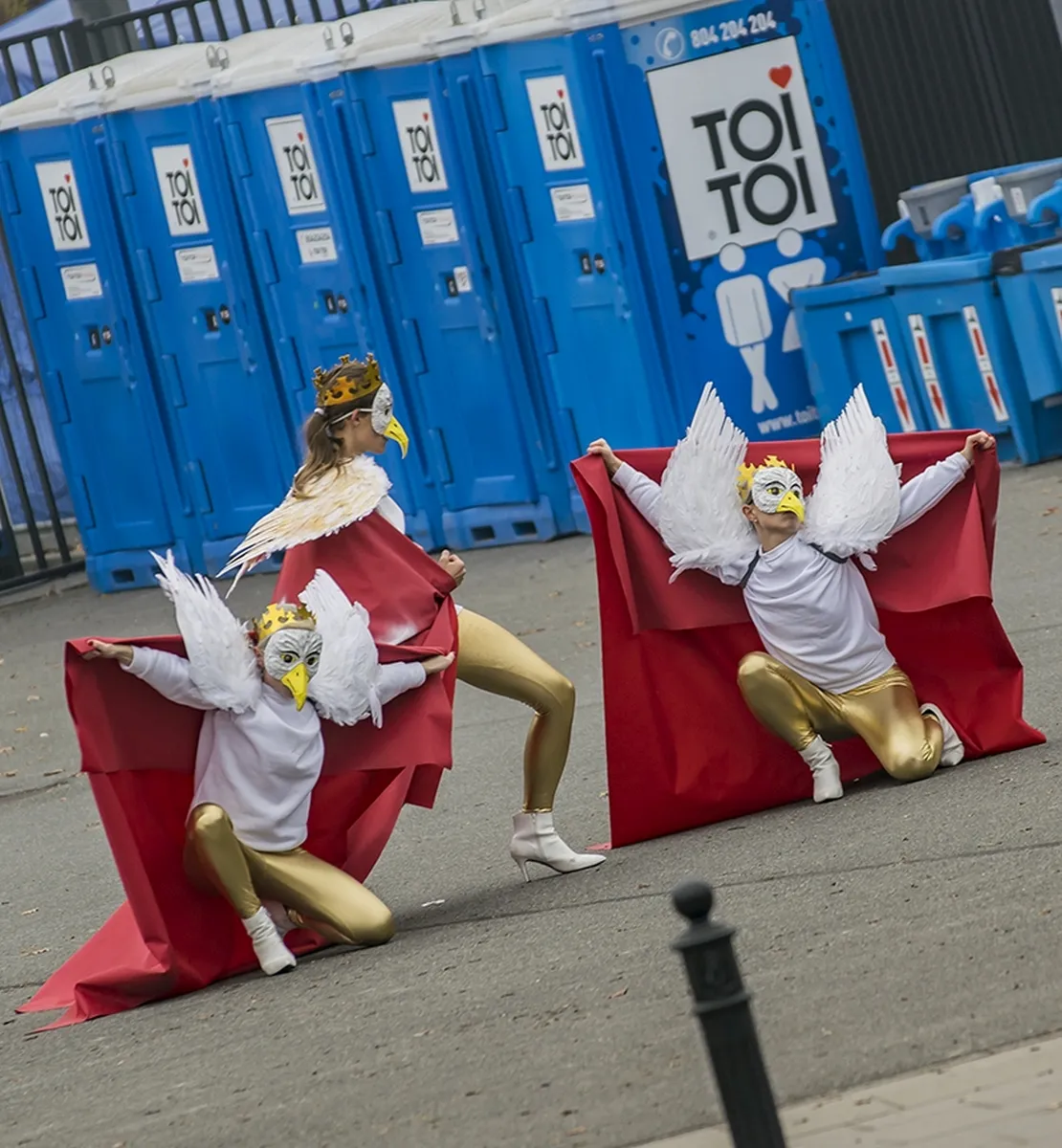 Three performers in bird masks and winged costumes posing before blue portable toilets